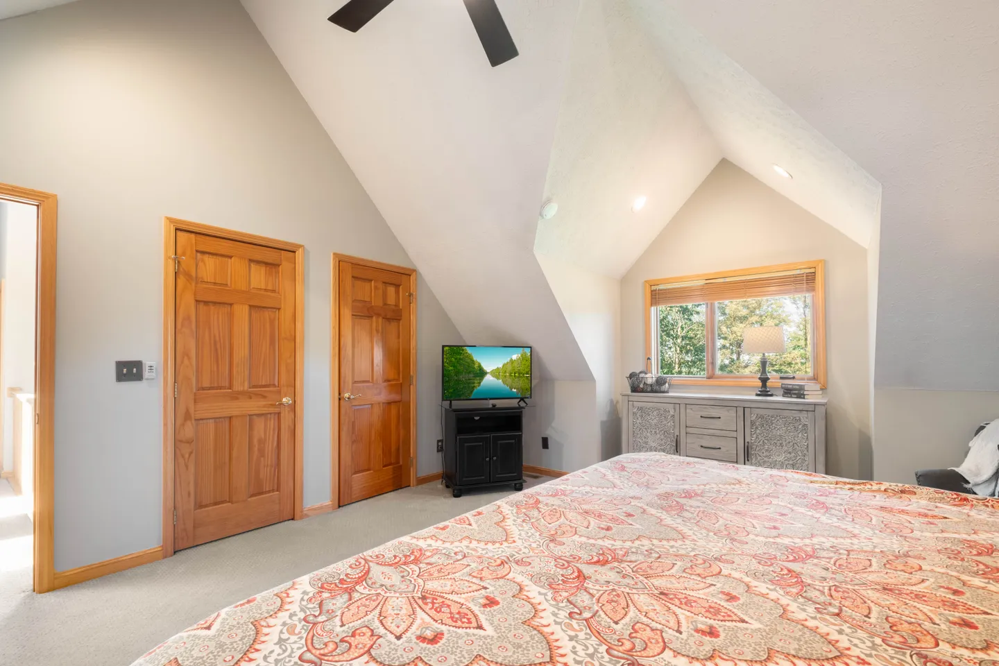 Bedroom with vaulted ceiling, patterned bedspread, television on a stand, wooden doors, and a window in an alcove.
