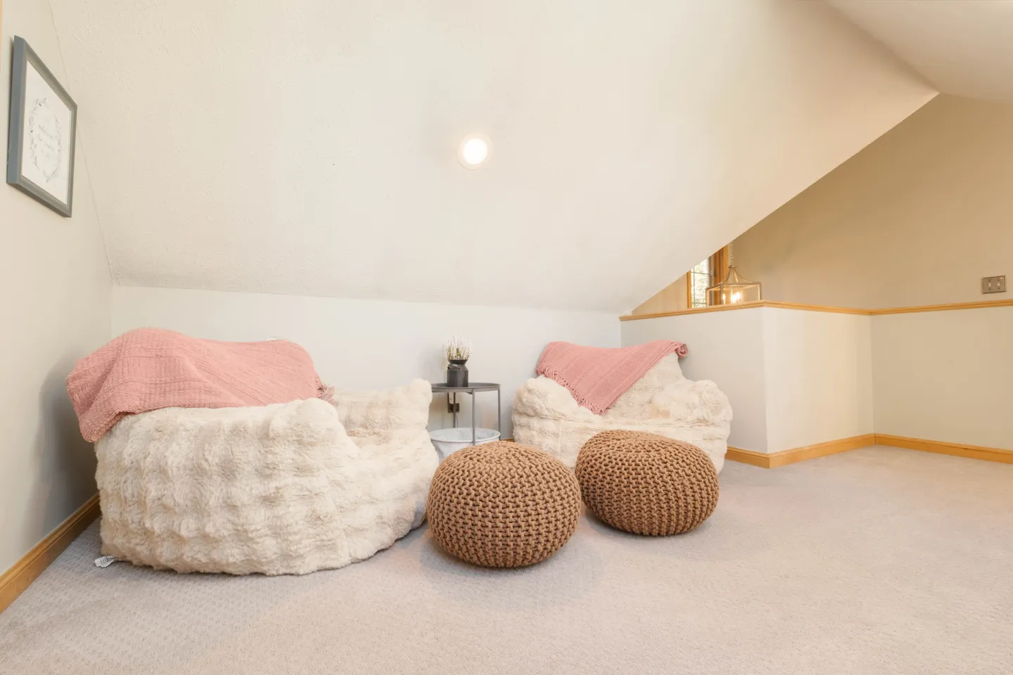 A cozy loft sitting nook featuring two plush beanbag chairs with pink throws, two knit poufs, and a small side table under a sloped ceiling.