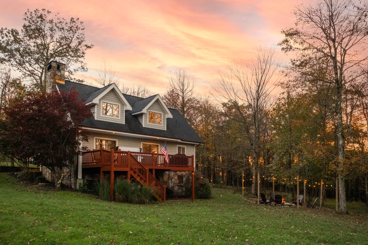 Exterior view of a multi-level home with an elevated wooden deck, stone foundation, and a backyard fire pit under a sunset sky.