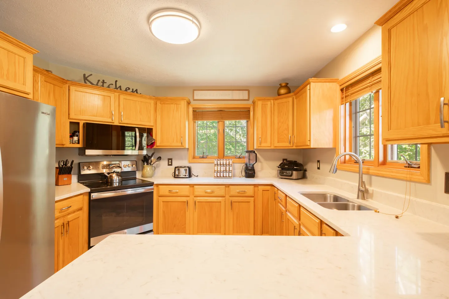 Kitchen featuring light wood cabinets, light-colored countertops, stainless steel appliances, and two windows.