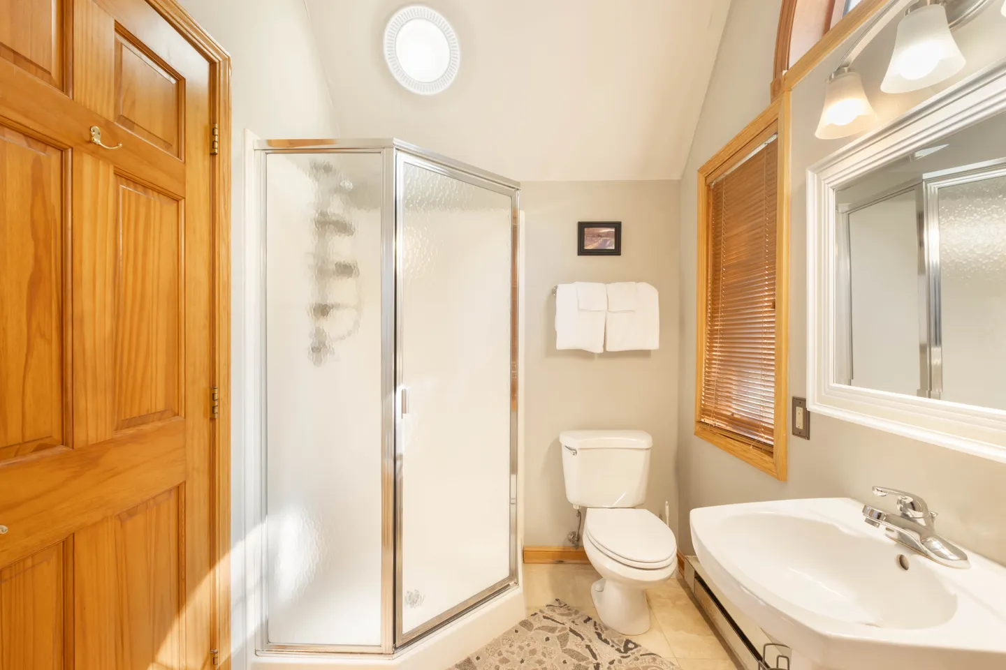 Bathroom with a corner walk-in shower, toilet, pedestal-style sink, and sloped ceiling with a round skylight.