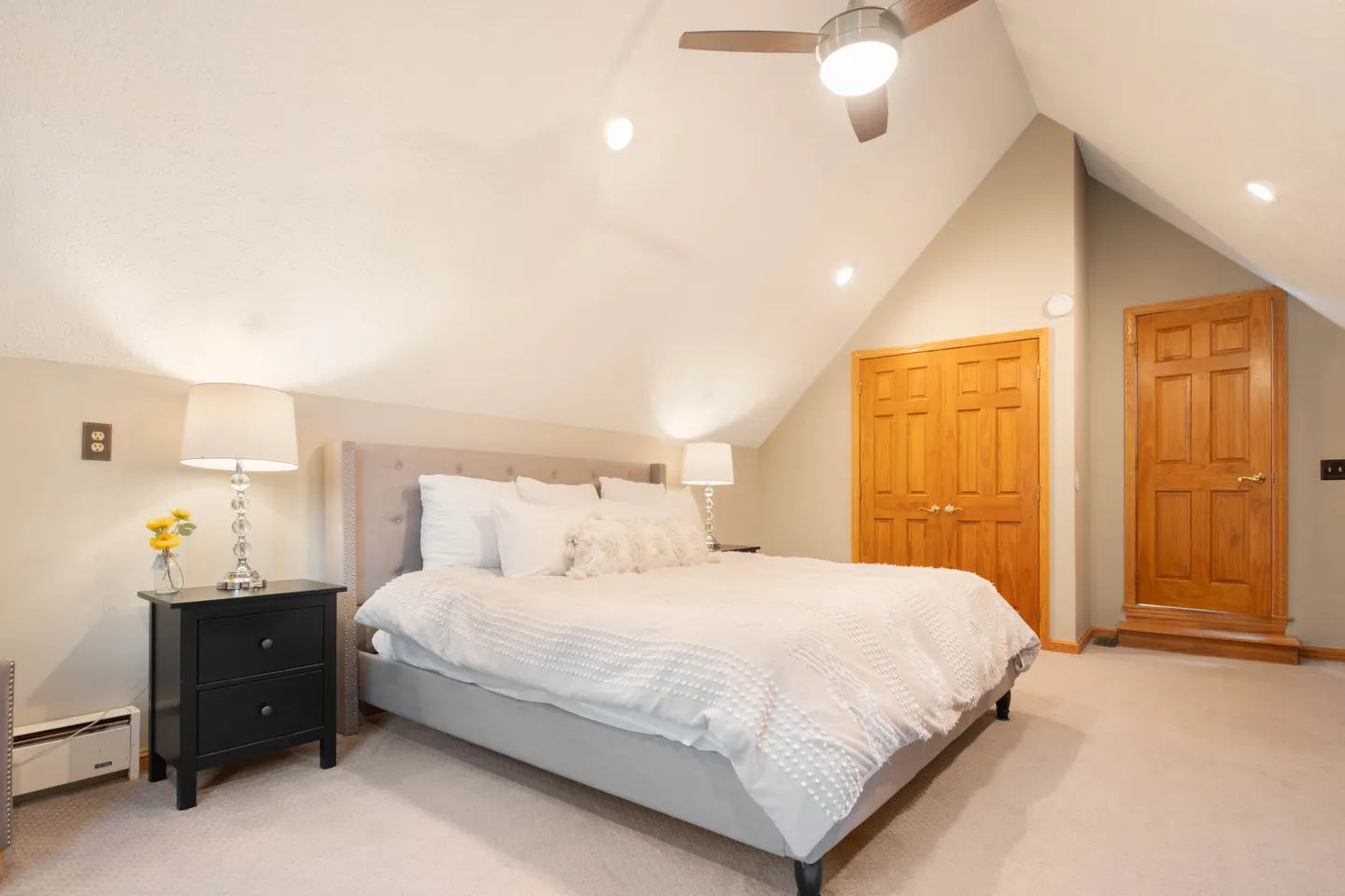 Bedroom with vaulted ceiling, upholstered bed, nightstands with lamps, ceiling fan, and wooden doors.