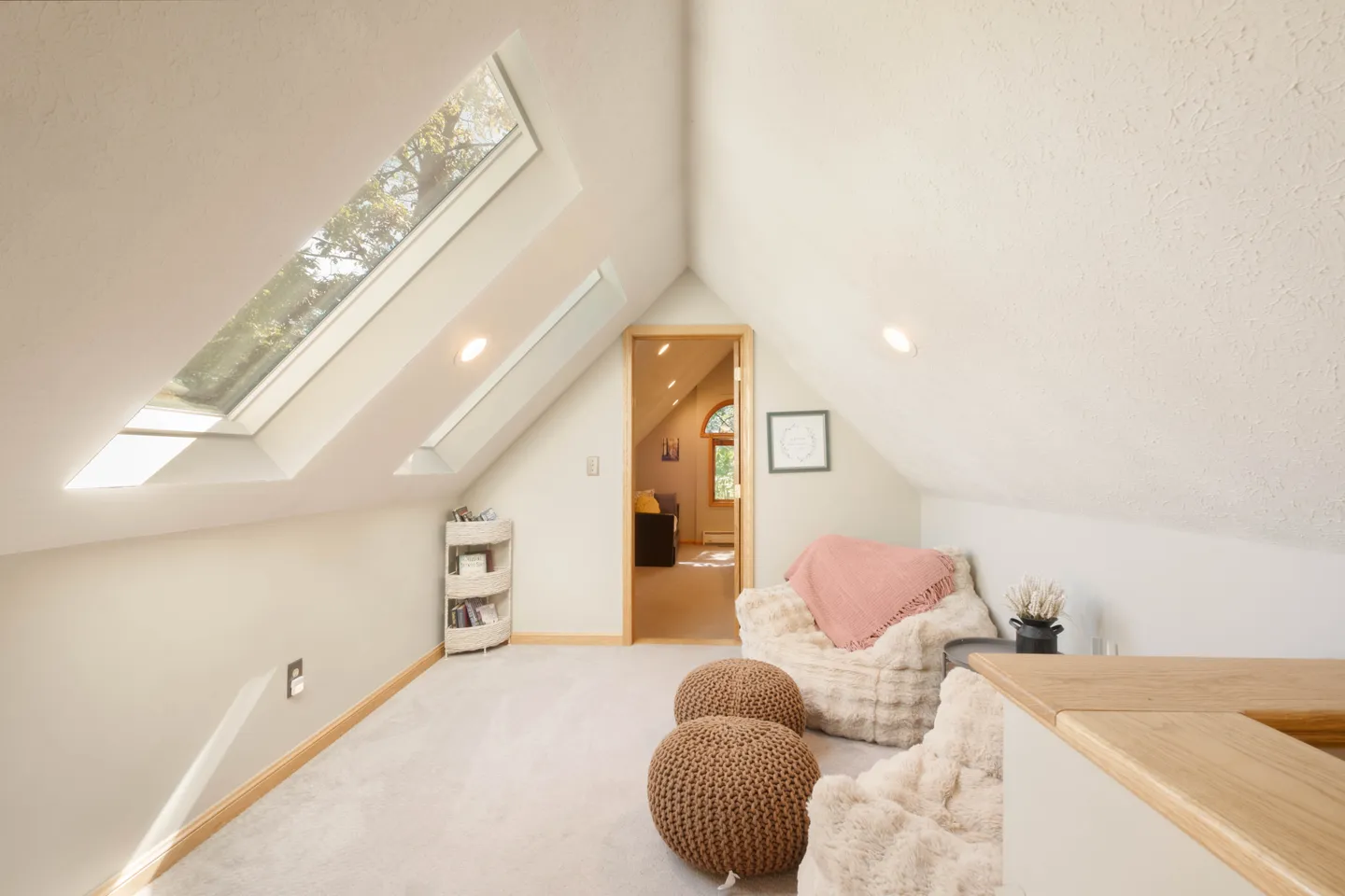 Bright attic seating area with angled ceiling, skylights, plush chairs, woven poufs, and an open doorway to another room.