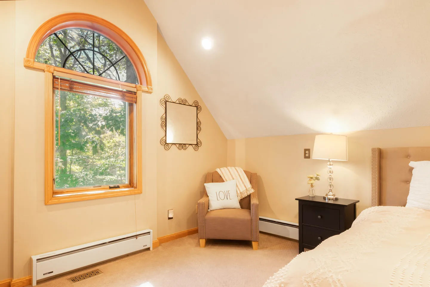 Bedroom with sloped ceiling, arched wood-framed window, armchair, nightstand, and edge of a bed.