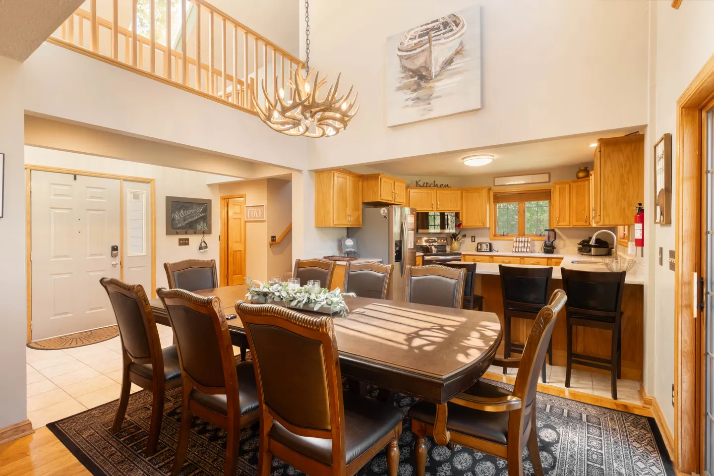 Dining area with a large wooden table, antler chandelier, and view into the adjacent kitchen under high ceilings.