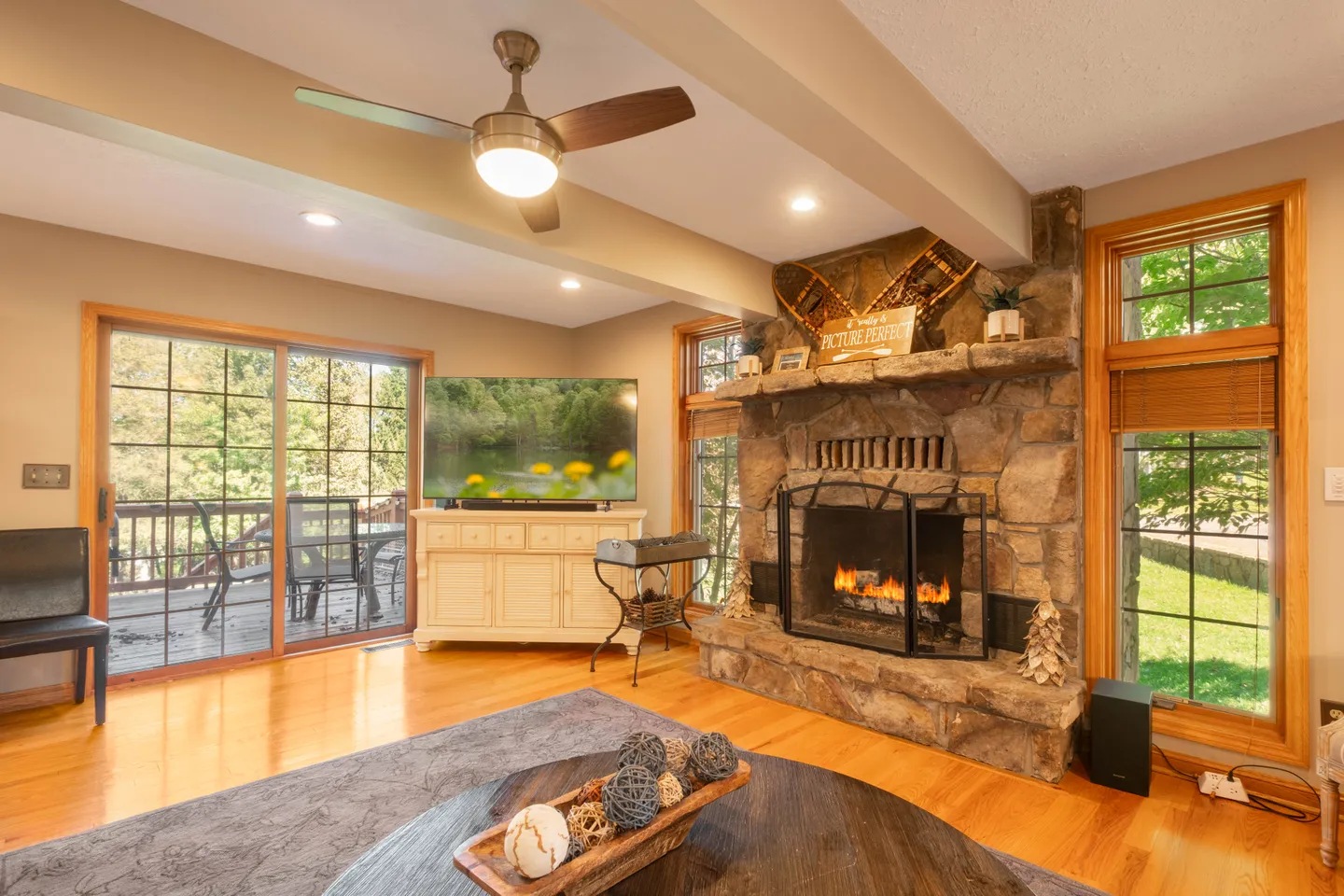 Living room with a stone fireplace, wood floors, flat-screen television, ceiling fan, and sliding glass doors leading to an outdoor deck.