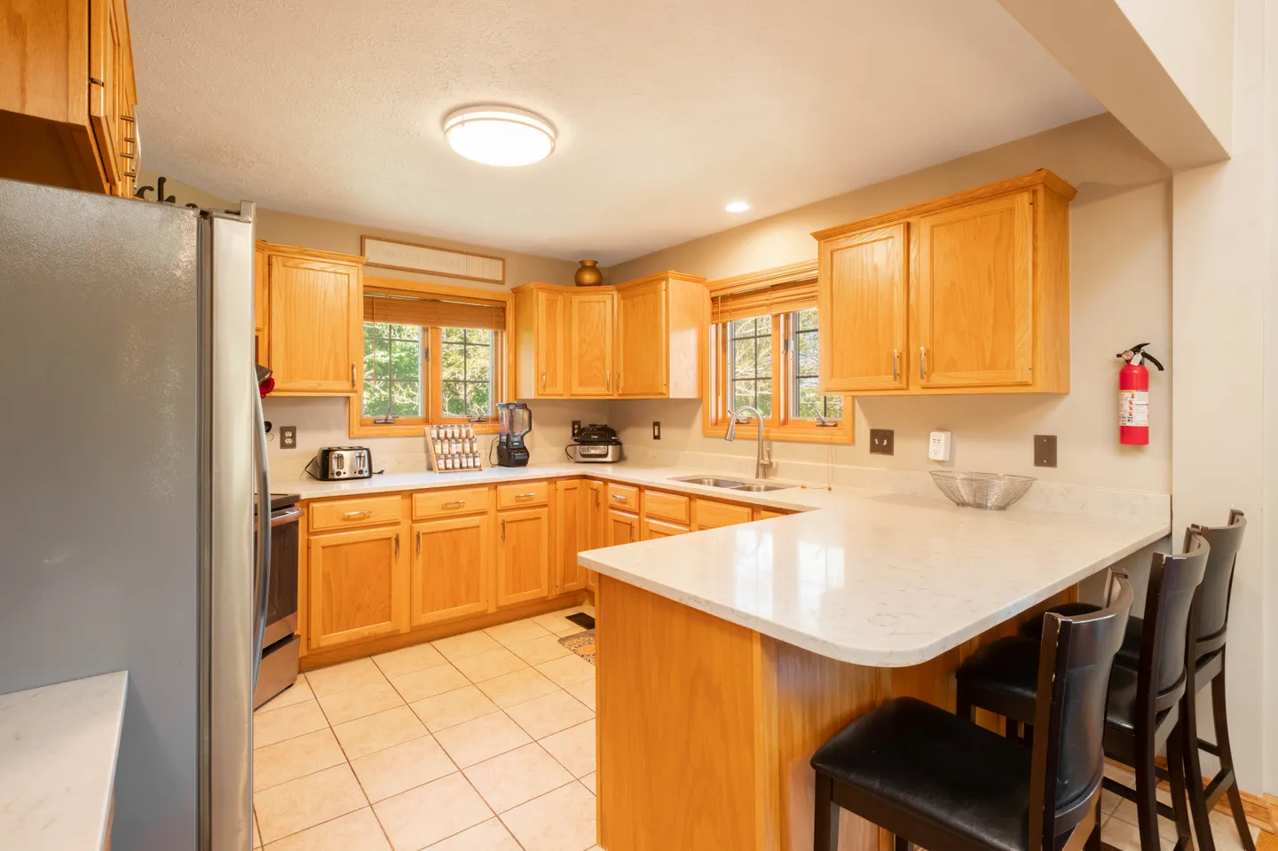 Kitchen with wooden cabinetry, white countertops, tiled flooring, windows, and an island counter with black barstools.