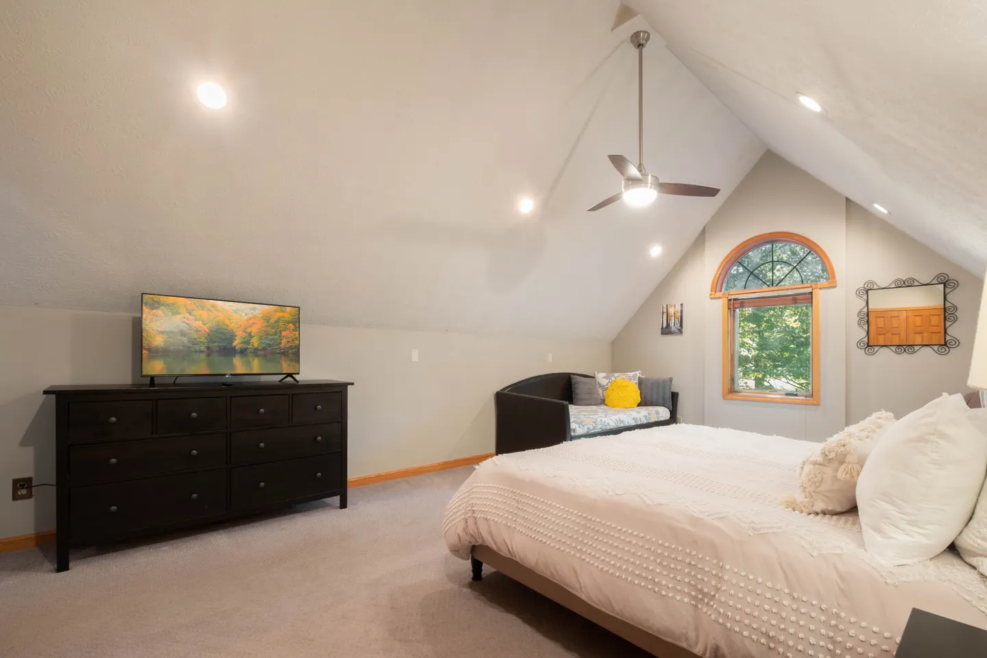 Attic-level bedroom with carpet, vaulted ceiling, large bed, daybed under a small arched window, and a dark wooden dresser with a TV.