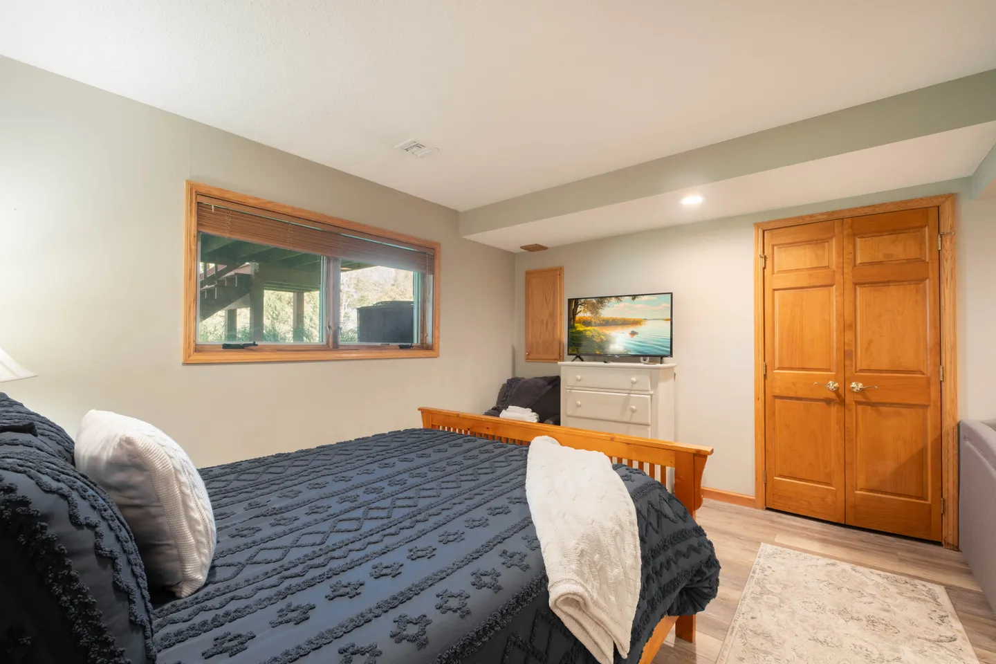 Bedroom with a wooden frame bed made up with a dark blue patterned quilt, a window looking outside, a dresser with a television, and a wooden double door.