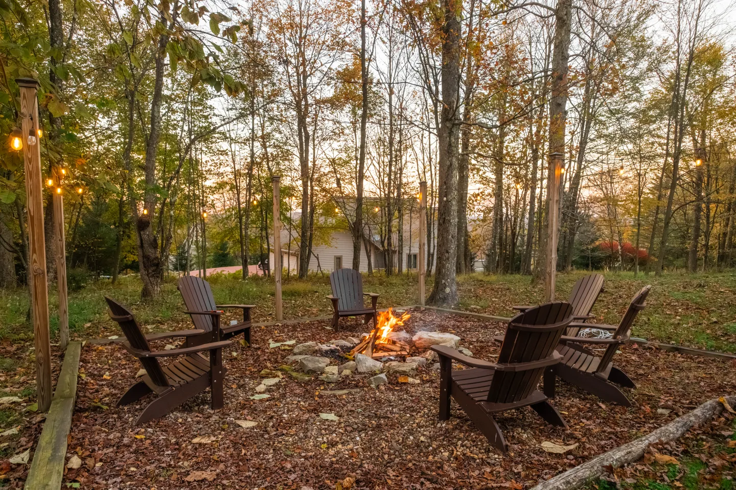 Outdoor fire pit surrounded by Adirondack chairs and string lights on wooden posts in a wooded yard.
