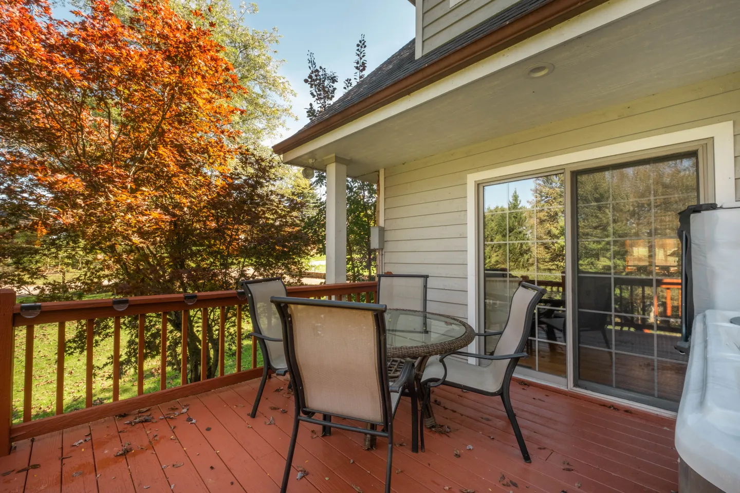 Outdoor wooden deck with a patio table, chairs, and sliding glass door leading into the house, surrounded by autumn trees.