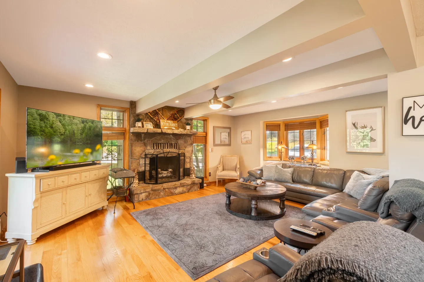 Living room with leather sectional sofa, stone fireplace, flat-screen television, and bay window