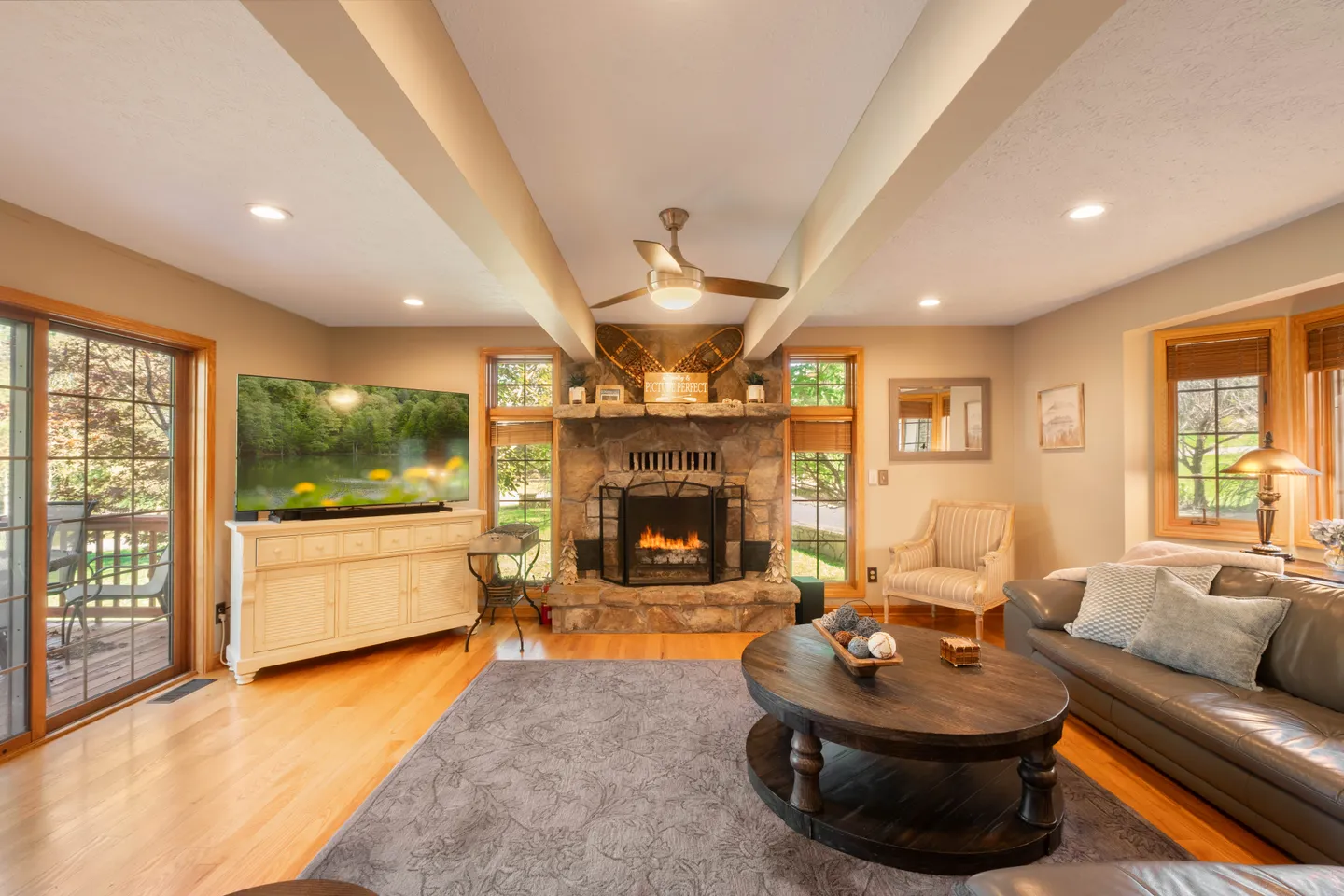 Living room with light wood floors, a central stone fireplace, a flat-screen TV on a sideboard, and sliding glass doors to a deck.
