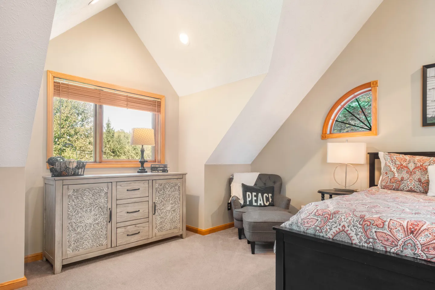 Bedroom with vaulted ceiling, patterned bedding on a dark frame bed, armchair with ottoman, wooden dresser, and an arched accent window.