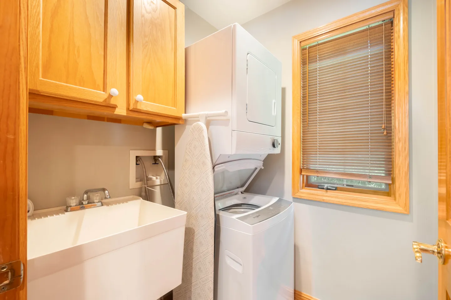 Laundry room with a stacked washer and dryer, deep utility sink, wooden upper cabinets, and an ironing board.