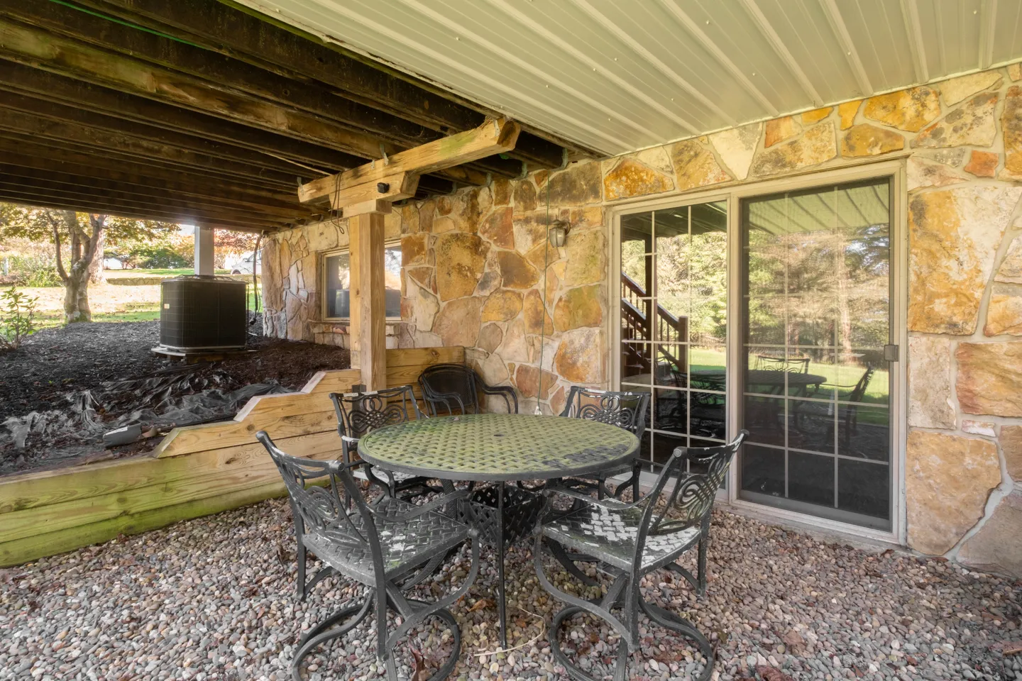 Covered gravel patio underneath an elevated deck, featuring a round table and metal chairs against a stone wall with sliding glass doors.