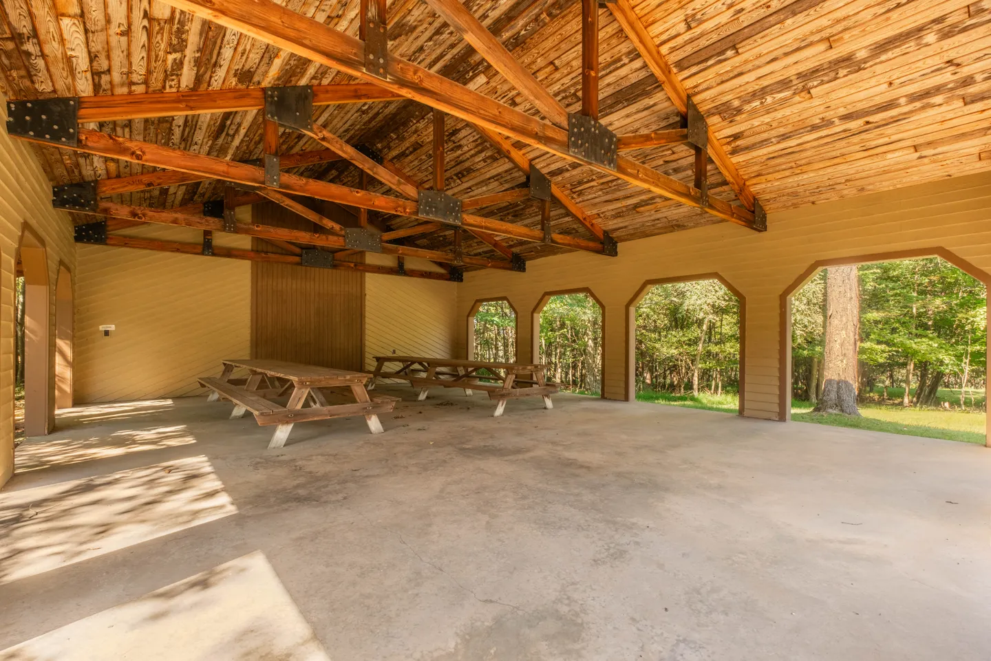 Open-air pavilion featuring exposed wooden roof trusses, concrete flooring, and picnic tables looking out to trees.