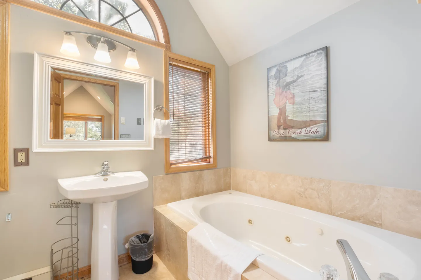 Bathroom featuring a pedestal sink under an arched window and a large jetted tub with tile surround.