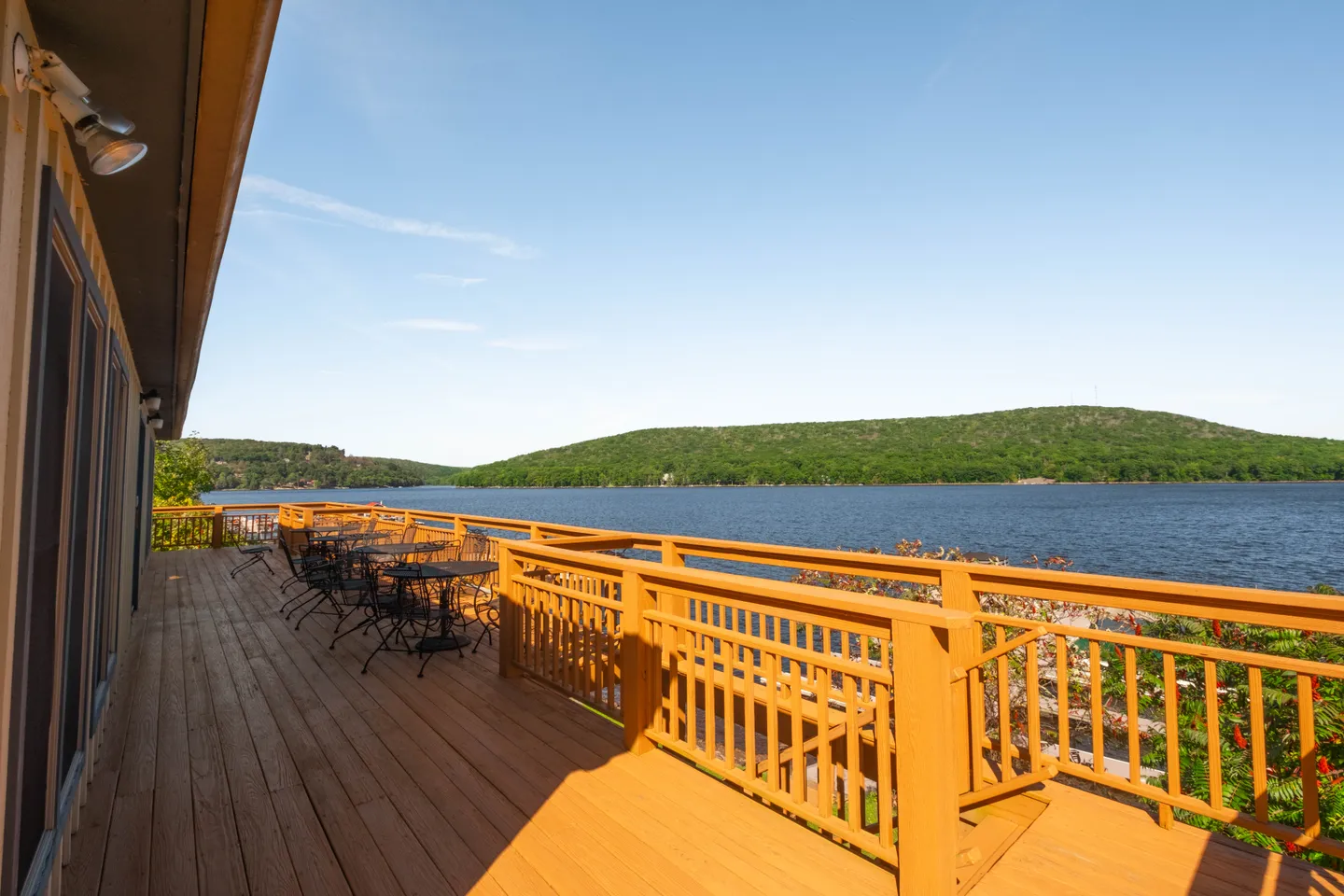 Spacious wooden deck with outdoor tables and chairs overlooking a wide lake and forested hills under a blue sky.