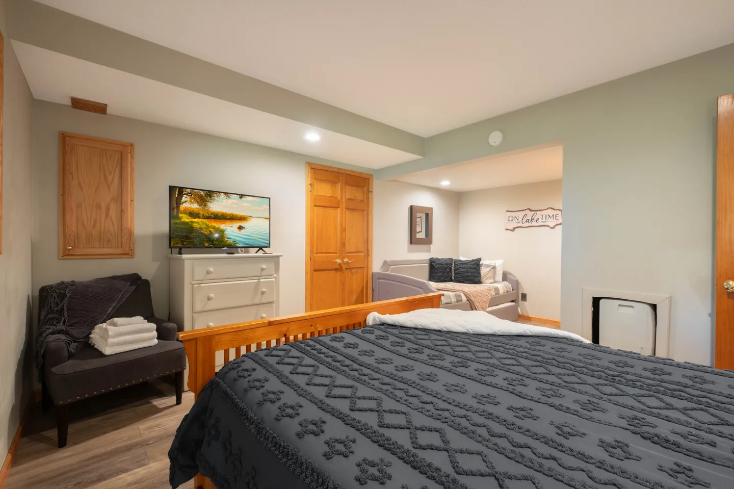 Bedroom featuring a main bed with a dark textured bedspread, an armchair with towels, a dresser with a TV, and an alcove with a daybed.