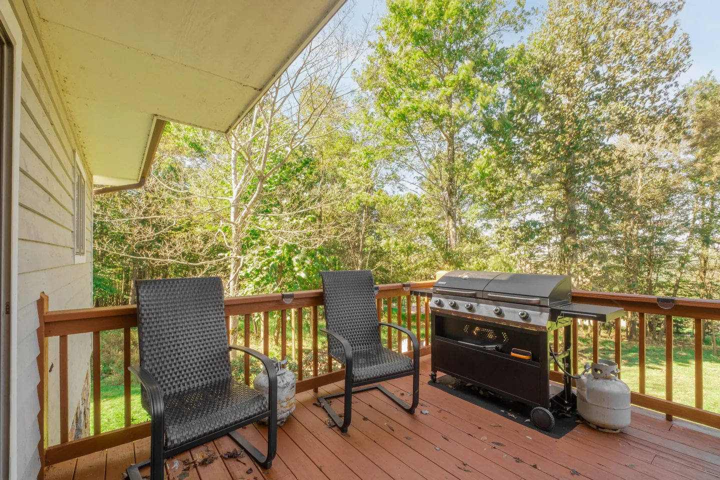 Outdoor deck with wooden railings, two dark woven chairs, and a gas grill overlooking lush green trees under a blue sky.