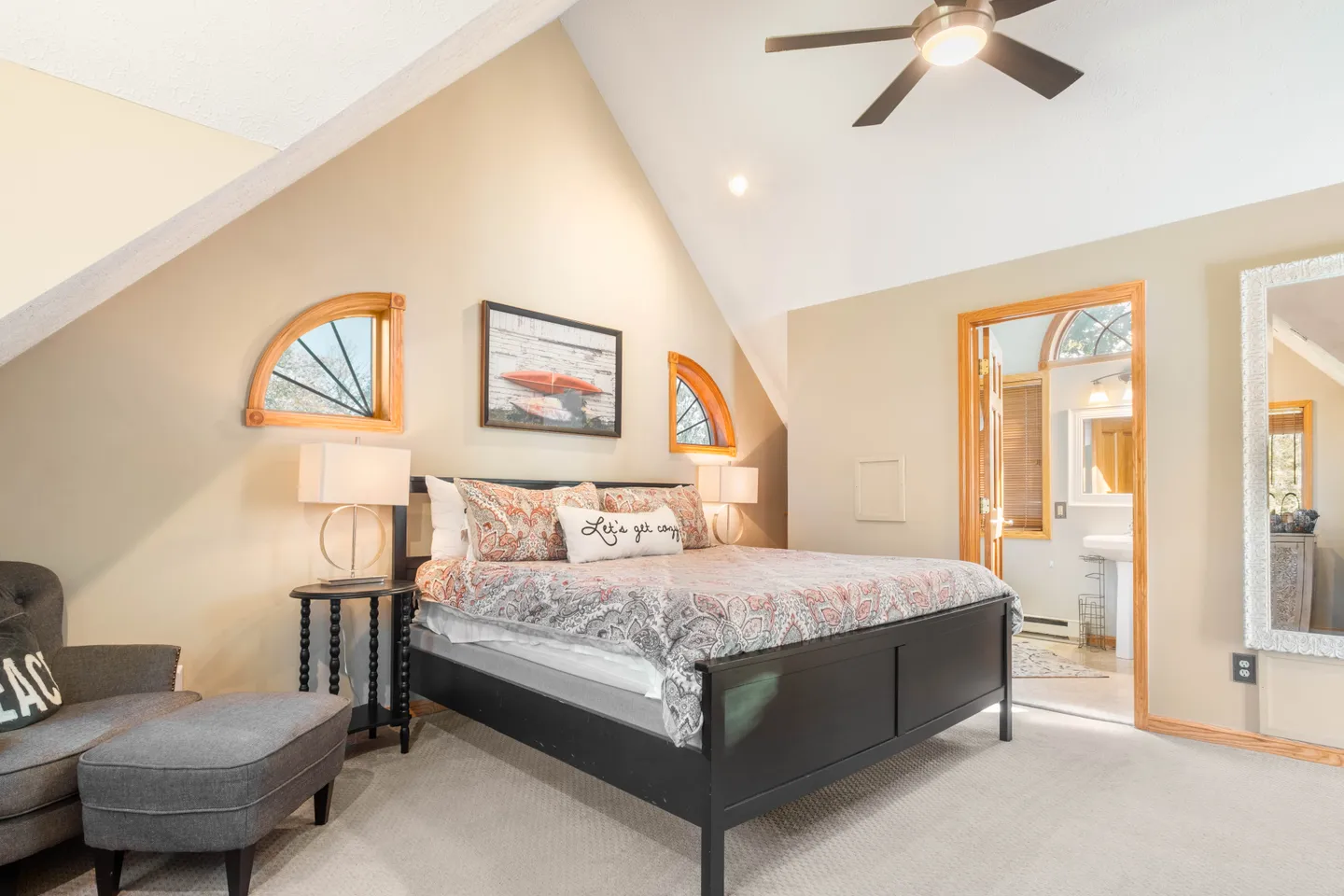 Bedroom featuring vaulted ceilings, a dark wood frame bed with patterned bedding, bedside tables with lamps, an armchair with an ottoman, and an en suite bathroom doorway.