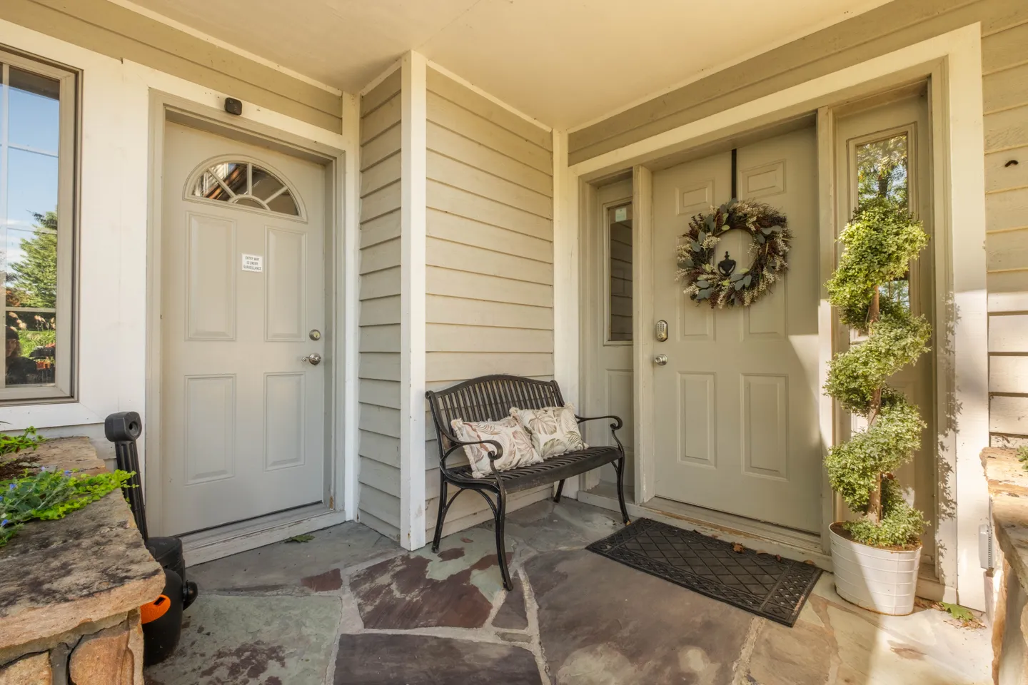 Covered stone front porch with two entry doors, a metal bench, and a spiral topiary.