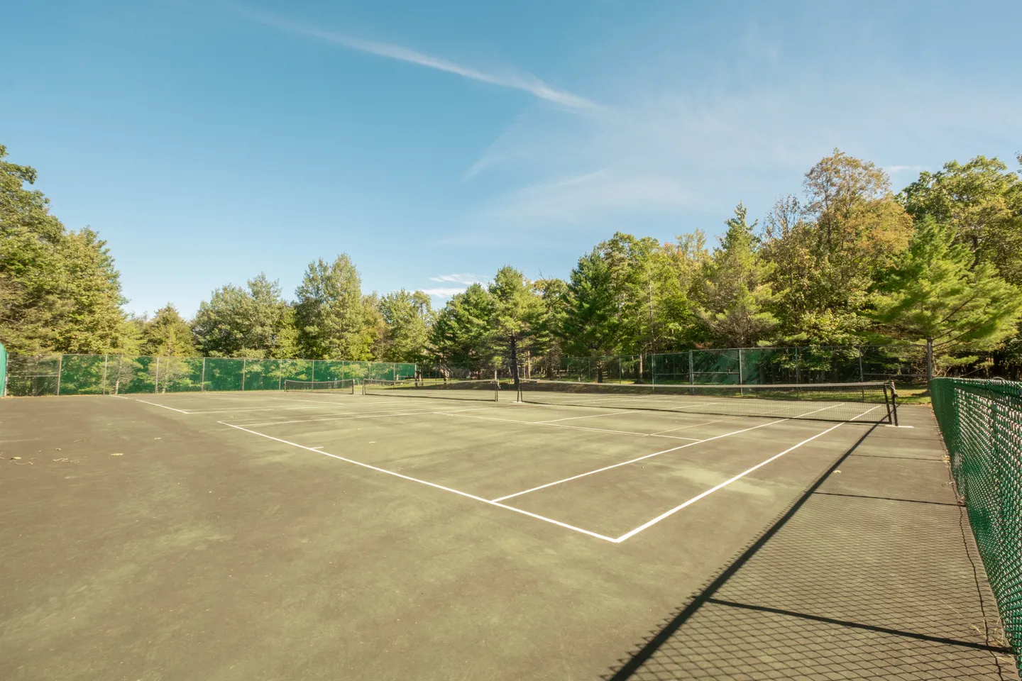 Outdoor tennis court enclosed by green fencing and surrounded by lush green trees beneath a blue sky.