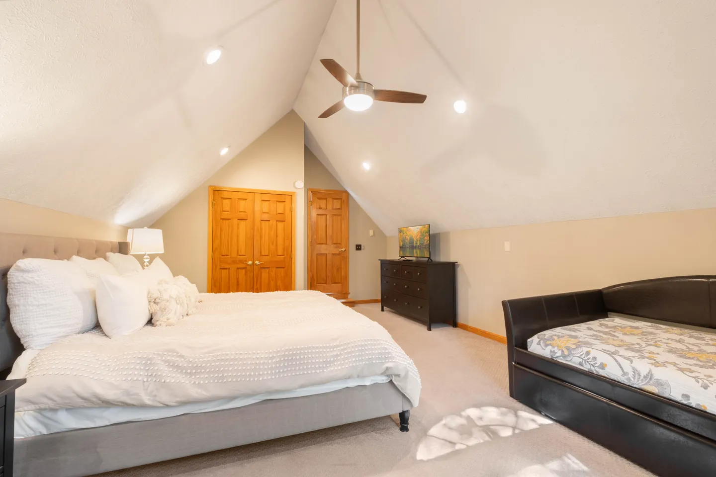 Attic-style bedroom with vaulted ceiling, large bed with white linens, daybed, ceiling fan, and dresser with TV.