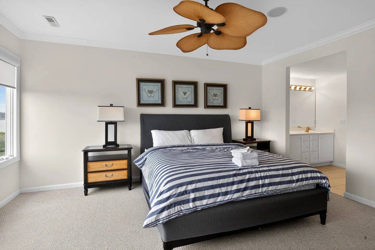 Bedroom with a dark upholstered bed, striped bedding, matching nightstands with lamps, a palm-leaf ceiling fan, and a view into an en-suite bathroom.