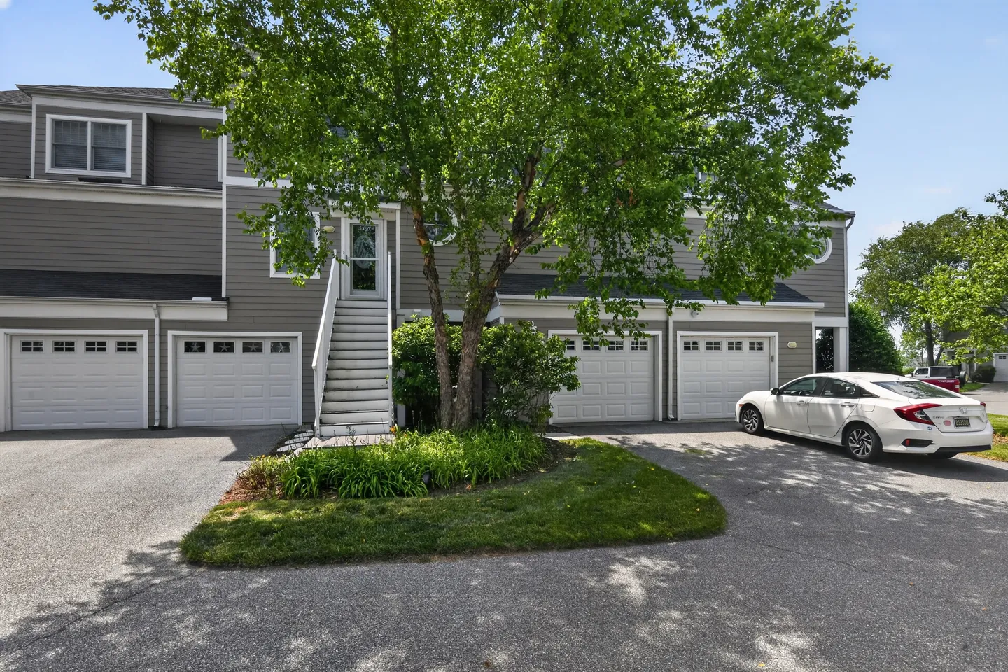 Exterior view of a multi-unit grey residential building with multiple white garage doors, an outdoor staircase, and a parked white sedan.