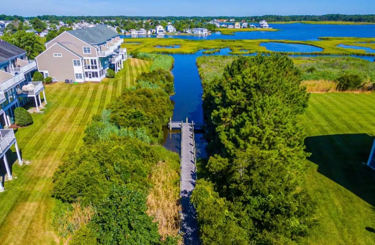Aerial view of a narrow canal and wooden dock flanked by lush green trees, striped lawns, and multi-story waterfront townhomes leading out to open bay waters.