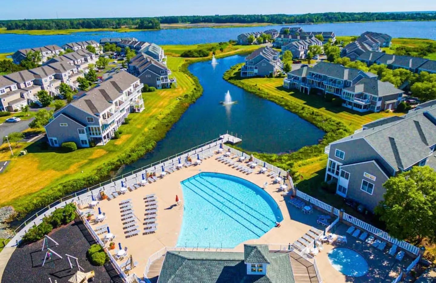 Aerial view of a community swimming pool complex alongside a pond with water fountains and nearby residential buildings.