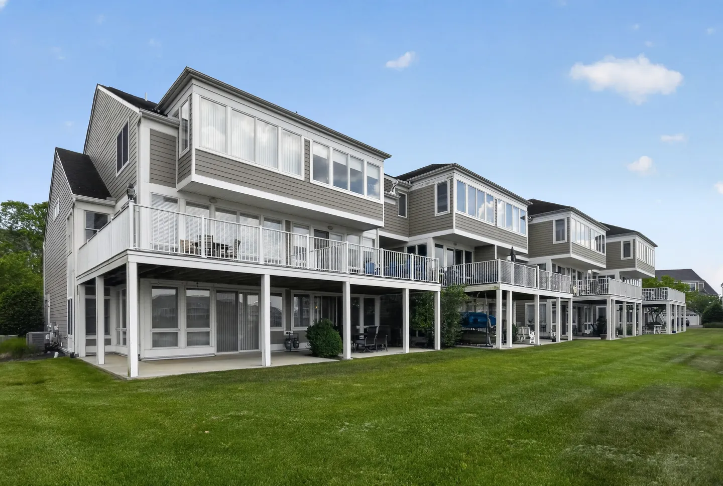 Rear exterior view of multi-story townhomes with raised decks and covered ground-floor patios bordering a green lawn.