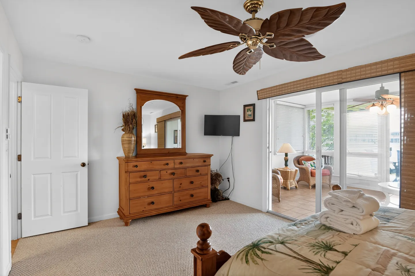 Bedroom featuring a wooden dresser with mirror, palm leaf ceiling fan, and sliding glass doors opening to an enclosed sunroom.