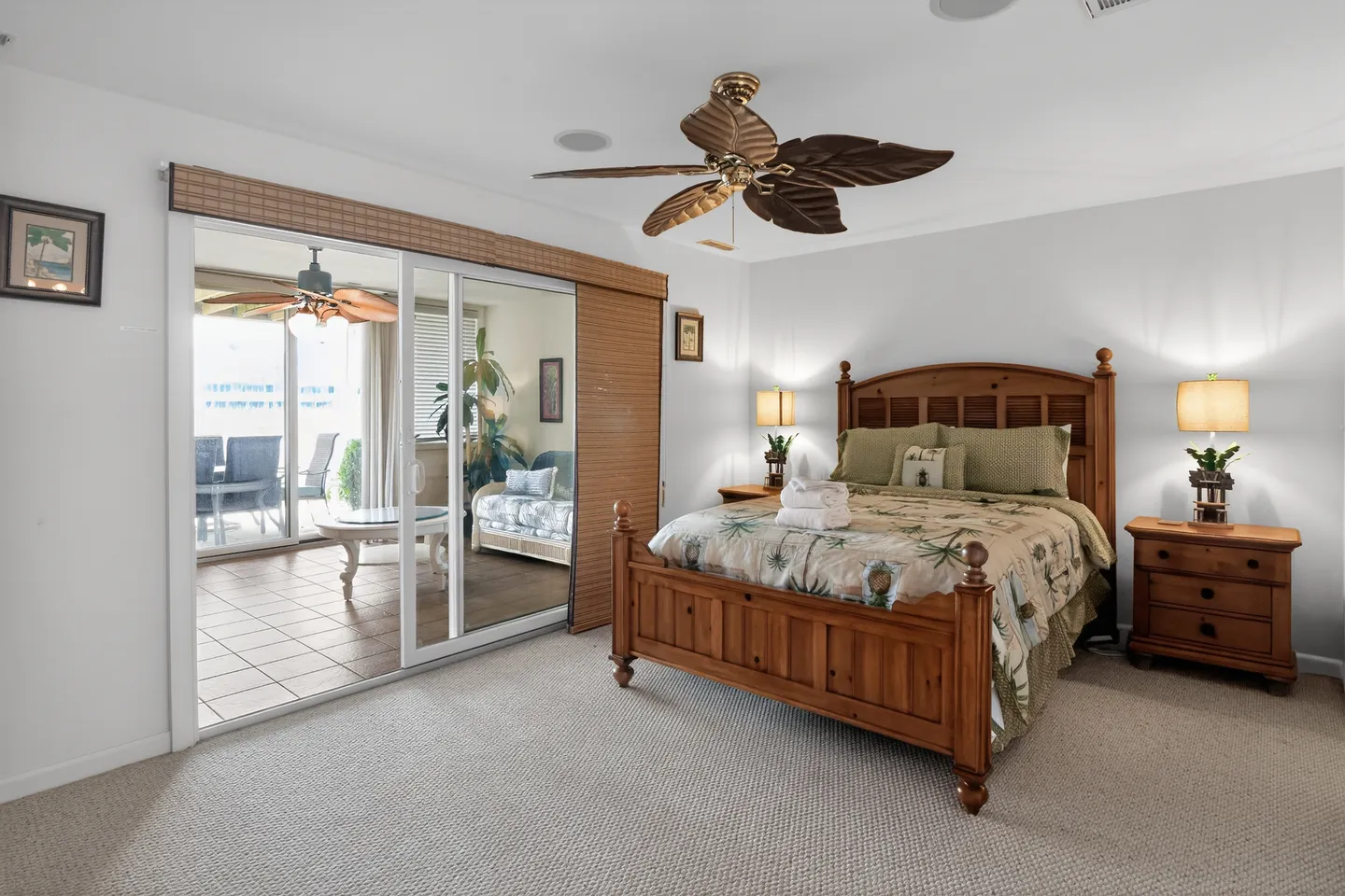 Bedroom with a wooden bed, patterned bedding, matching nightstands with lamps, a ceiling fan with palm leaf blades, and sliding glass doors leading to an enclosed sunroom.