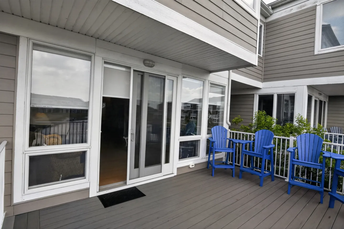 Outdoor deck with composite planking, blue chairs, white railings, and sliding glass door entrance beneath a balcony overhang.