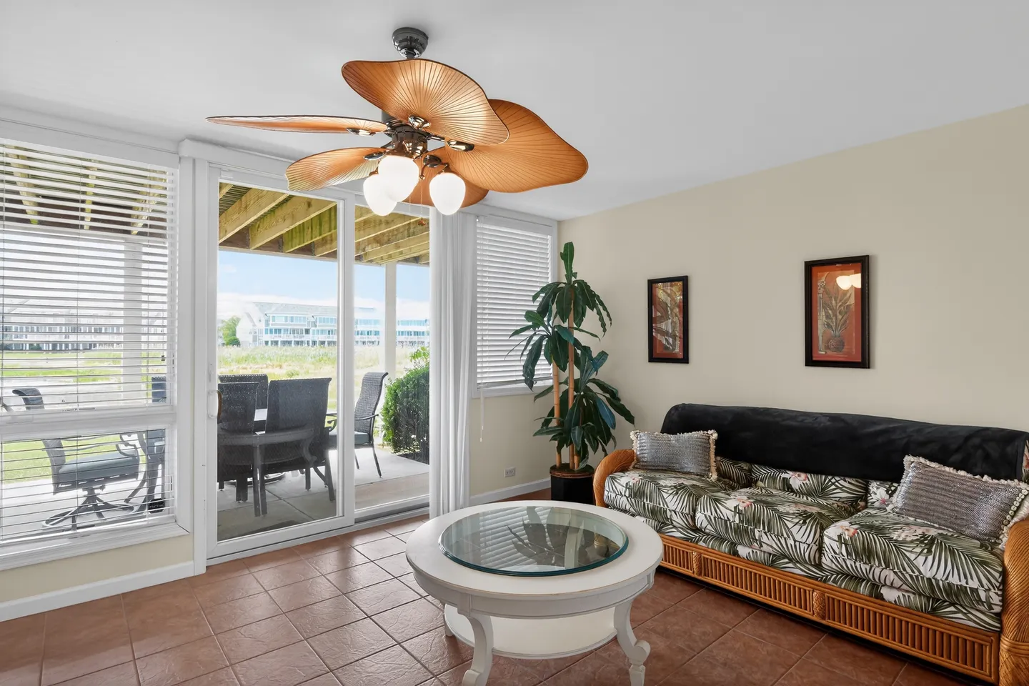 Sunroom with tiled floor, palm leaf ceiling fan, wicker sofa with tropical print cushions, round coffee table, and sliding glass doors leading to a patio.