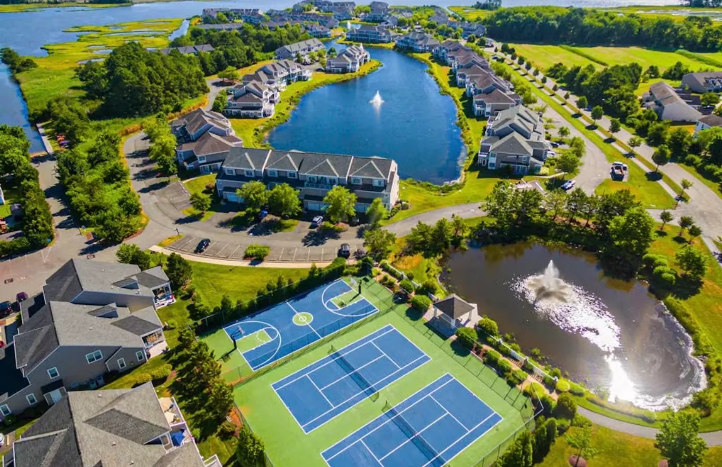 Aerial view of a residential community featuring ponds with fountains, tennis courts, a basketball court, and townhomes.