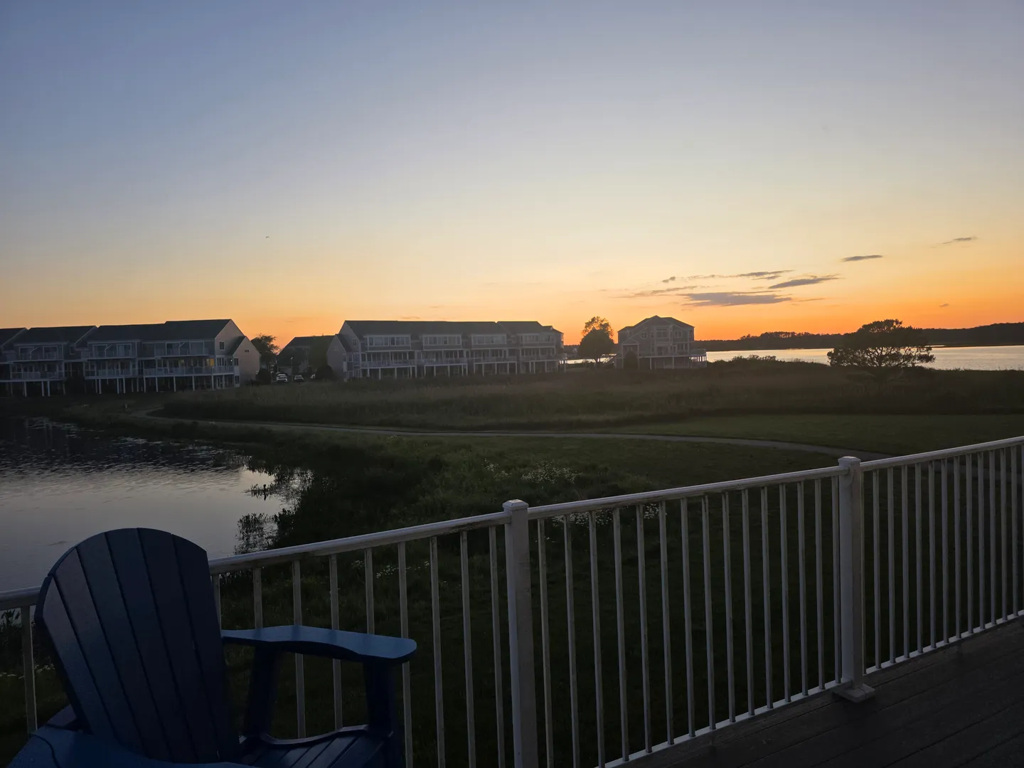 Deck view at sunset with an Adirondack chair overlooking a pond, grassy field, and neighboring buildings.