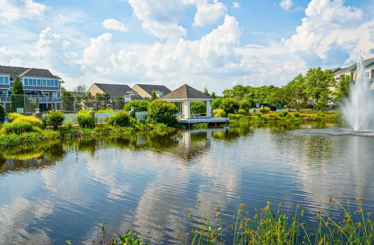 Outdoor pond with a water fountain and a gazebo along the grassy shoreline, with townhomes in the background under a blue sky.
