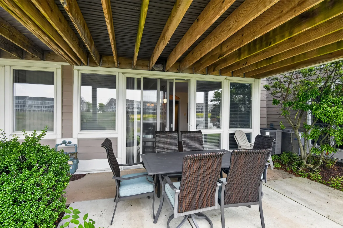 Covered concrete patio with an outdoor dining table and wicker chairs beneath an elevated wooden deck structure.