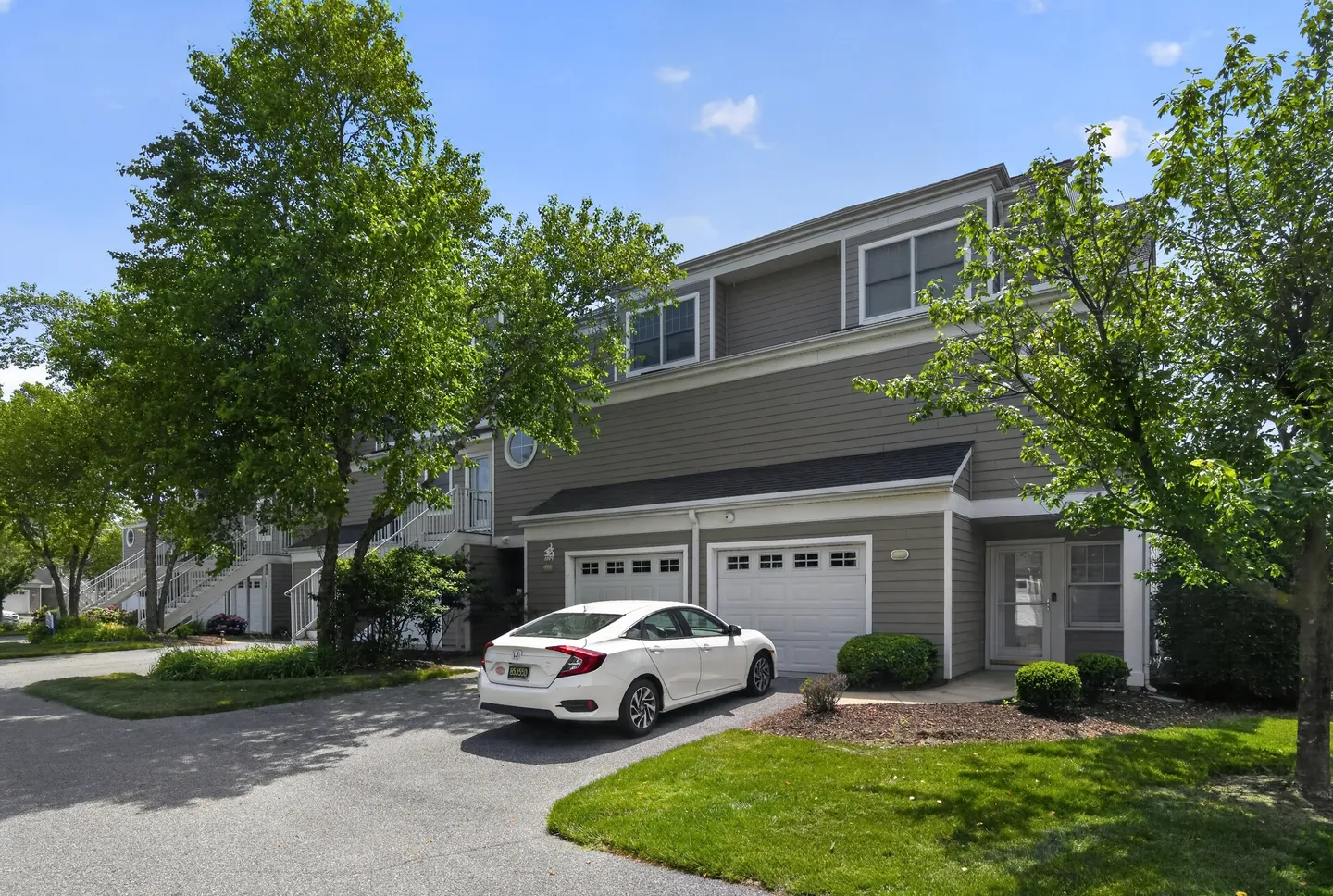 Exterior view of a multi-story gray townhouse with garage doors, an asphalt driveway, and a white sedan parked in front.
