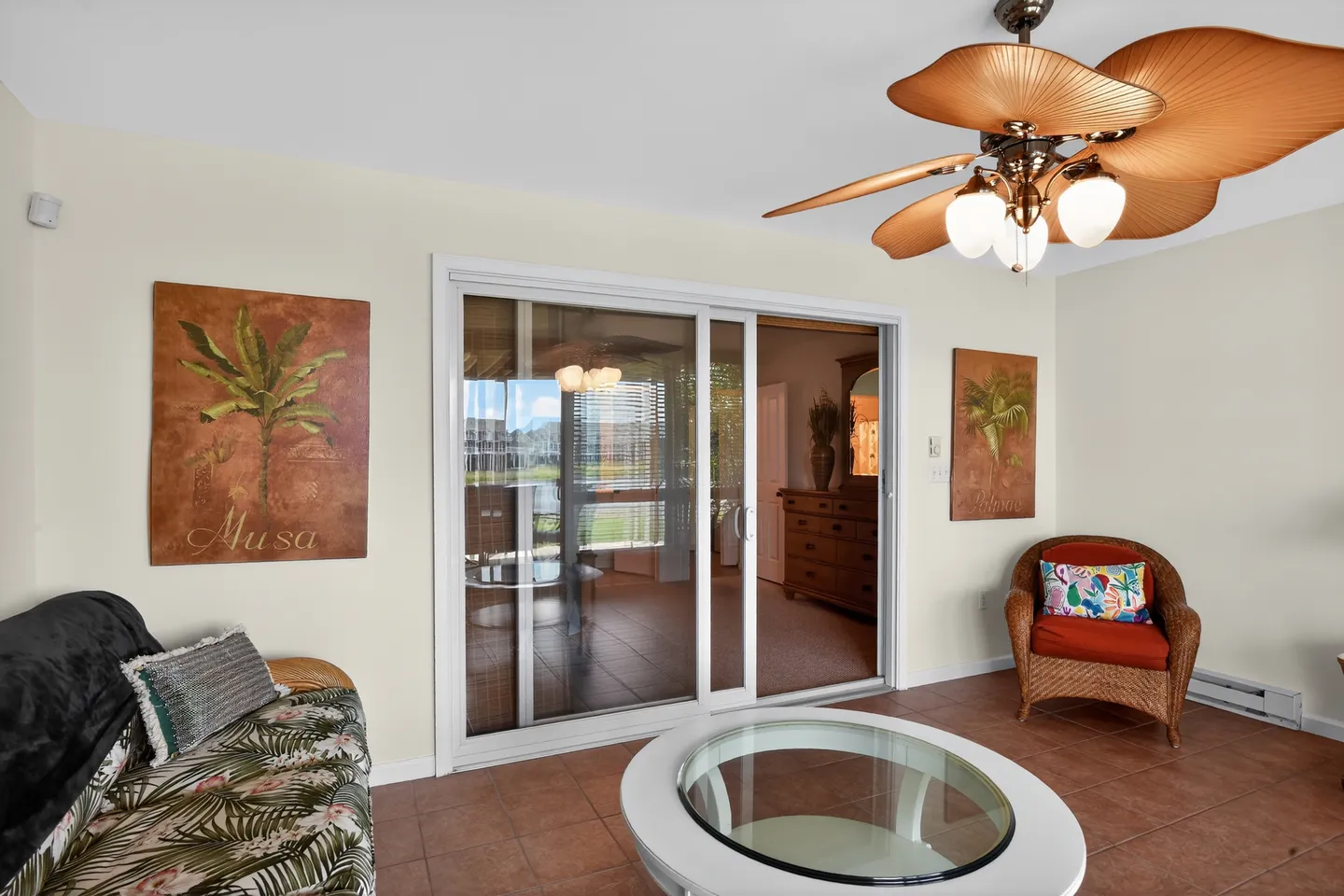 Sunroom with tiled floor, palm leaf ceiling fan, wicker armchair, sofa with tropical print cushions, and sliding glass doors to the interior.