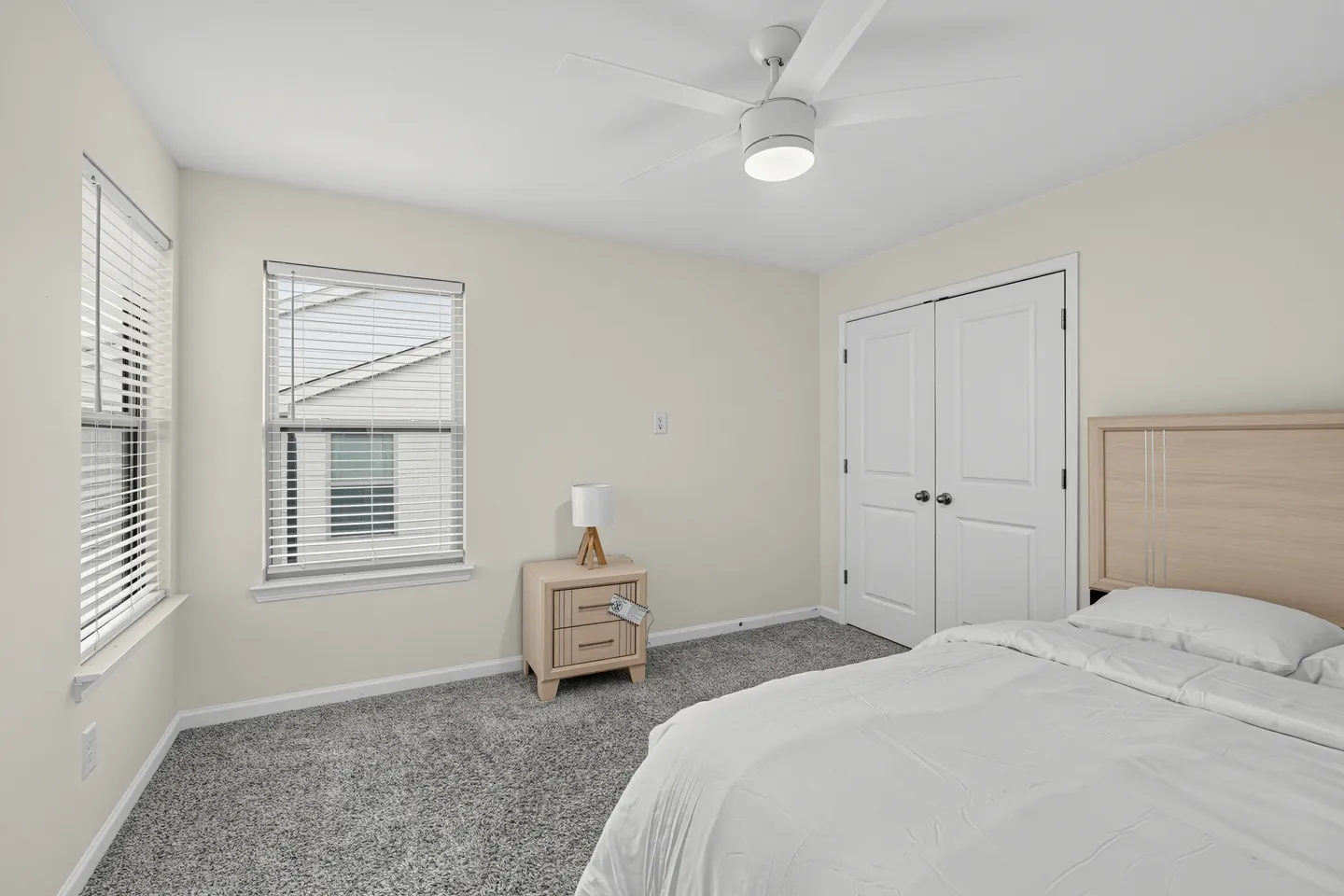 Bedroom with carpeted floor, ceiling fan, white closet doors, a bed, nightstand, and two windows with horizontal blinds.
